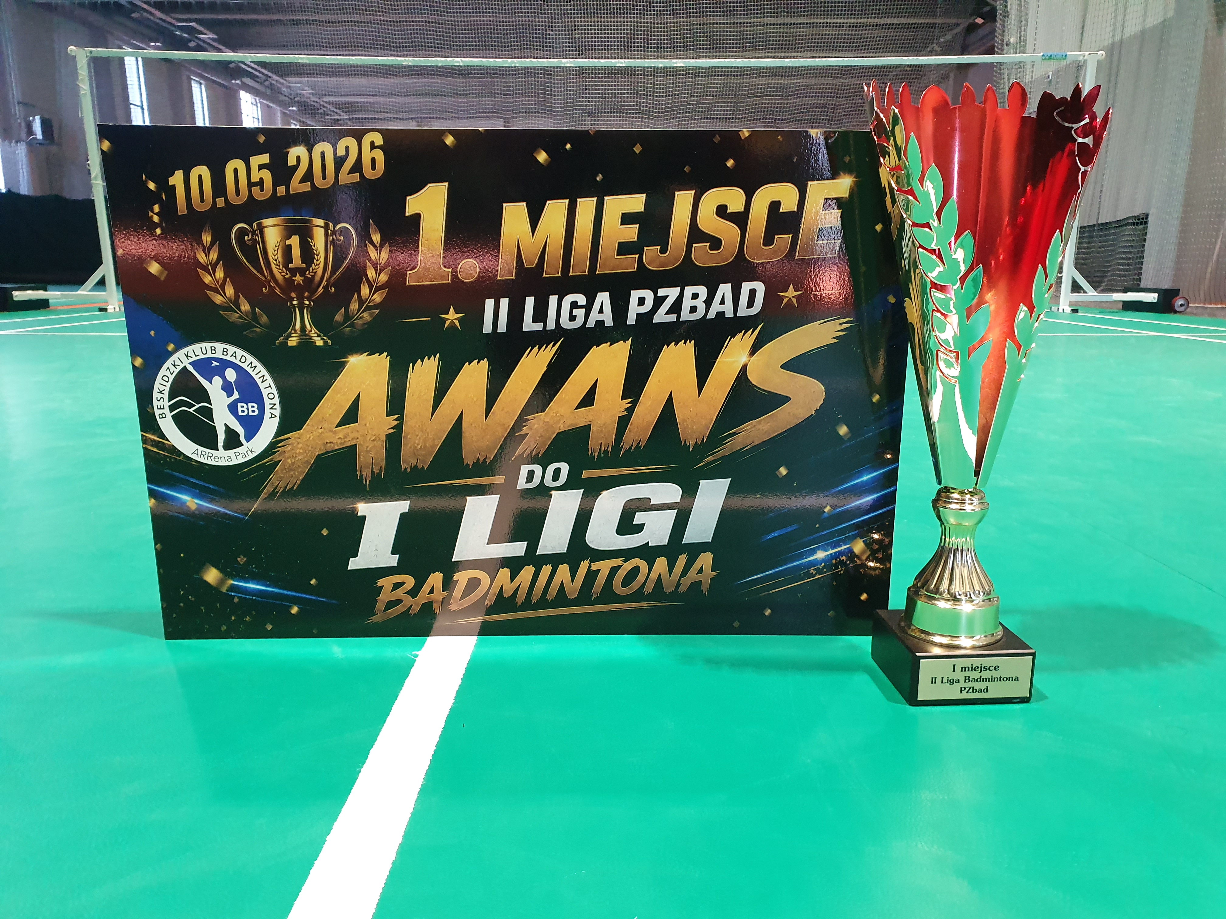 Puchar ustawiony obok tablicy z napisem „1. miejsce, II Liga PZBad, awans do I ligi badmintona”. Zdjęcie wykonano na korcie badmintonowym.